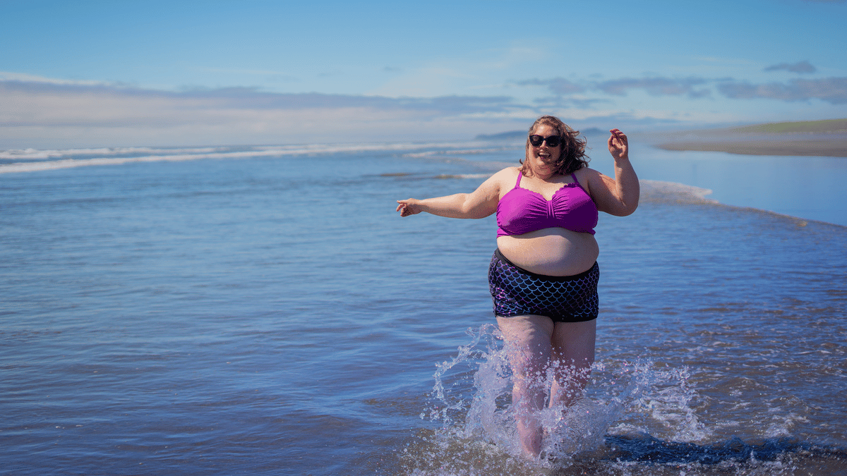 Woman in a larger body skipping along the water's edge at a beach, laughing and smiling